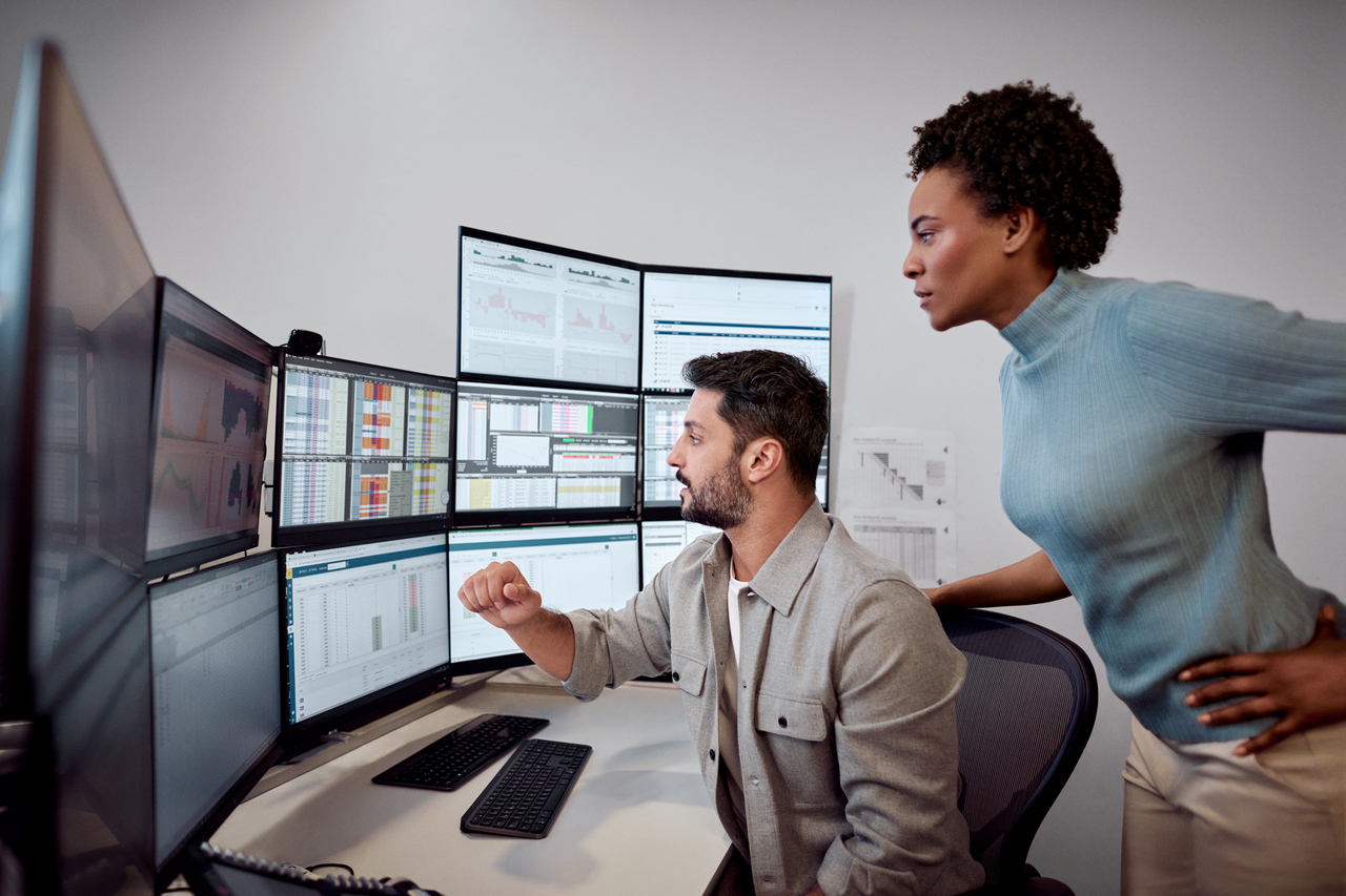 Ein Mann sitzt an einem Schreibtisch und zeigt einer Frau, die hinter ihm steht etwas auf seinen Bildschirmen.