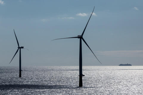 Zwei Windräder mitten im Meer. Im Hintergrund ist ein Schiff zu sehen.