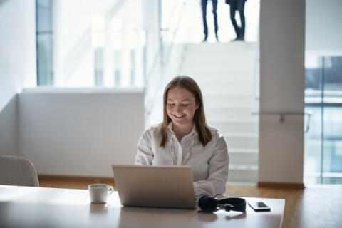 Eine Frau sitzt lächelnd vor ihrem Laptop in einer Büroumgebung