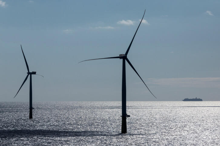 Zwei Windräder mitten im Meer. Im Hintergrund ist ein Schiff zu sehen.