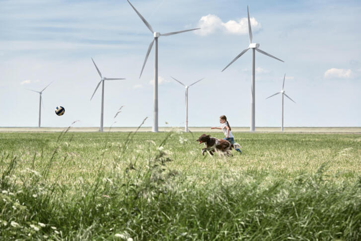 Eine grüne Wiese, auf der ein Mädchen mit ihrem Hund hinter einem Ball herläuft. Im Hintergrund Windräder.