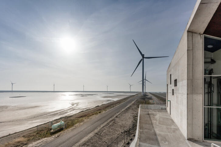 Ein Industriegebäude am Rand des Ufers. Am Horizont mehrere Windräder im Wasser sowie ein Windrad an Land.