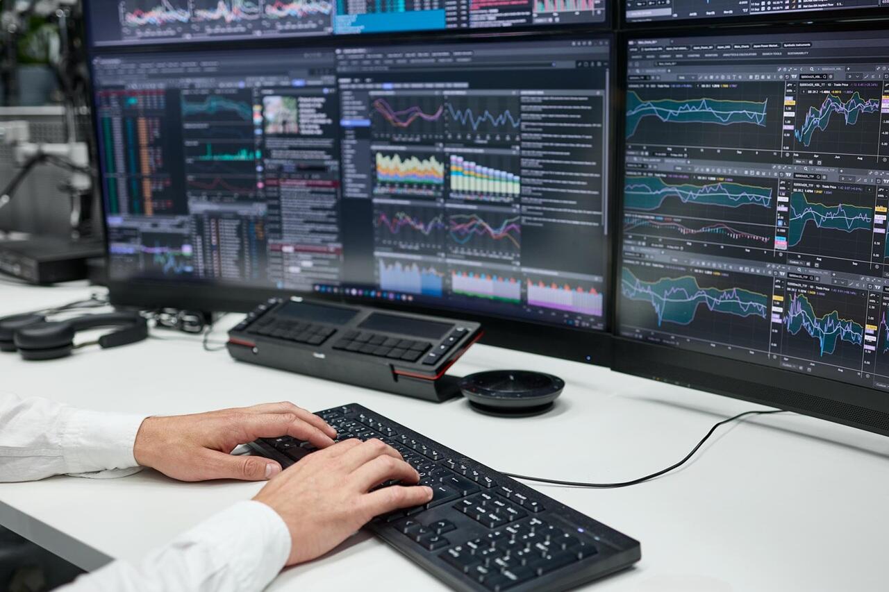 Hände eines Mannes auf der Tastatur vor mehreren Bildschirmen mit Graphen drauf