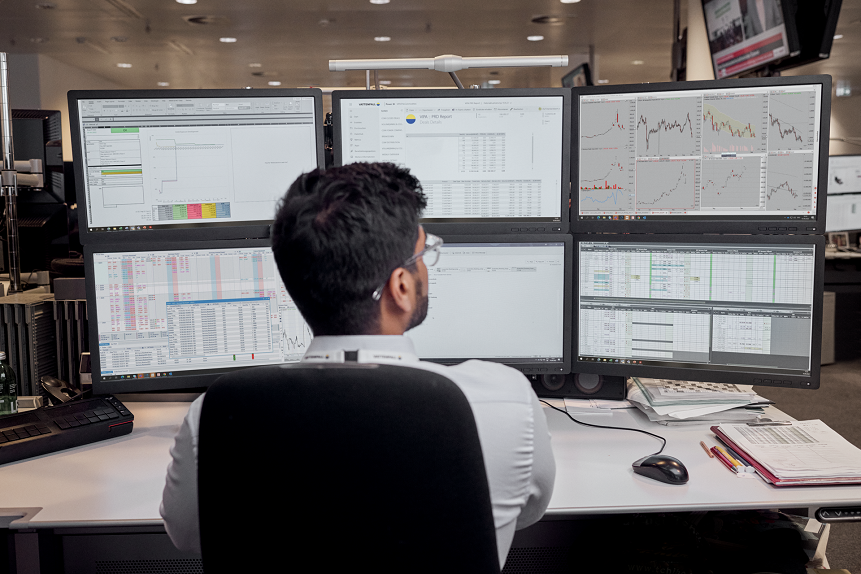 Ein Mann sitzt an einem Schreibtisch mit 6 Bildschirmen, auf denen unterschiedliche Graphen abgebildet sind.