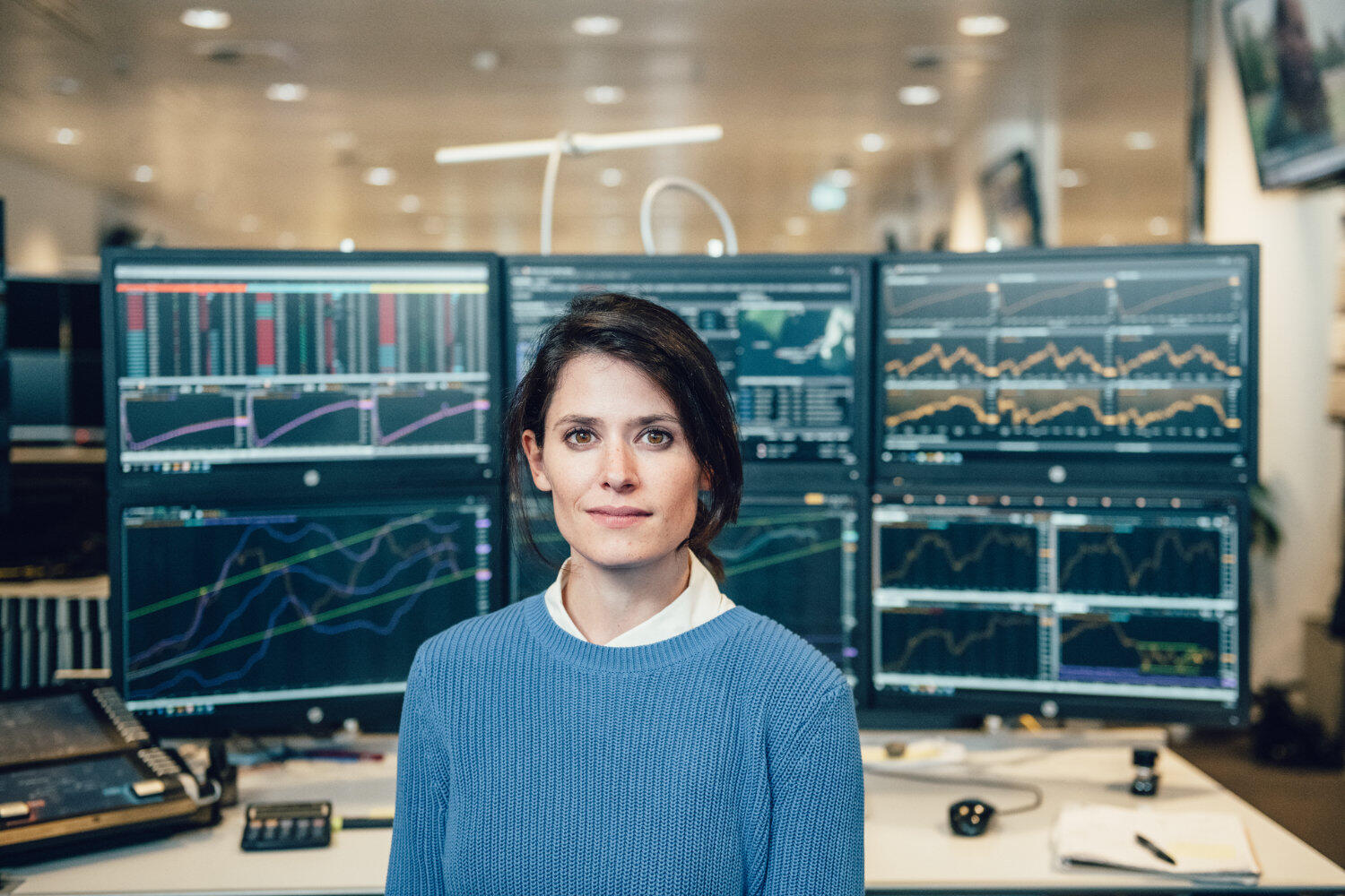 Eine Frau steht vor einem Schreibtisch mit 6 Bildschirmen, auf denen Graphen abgebildet sind.