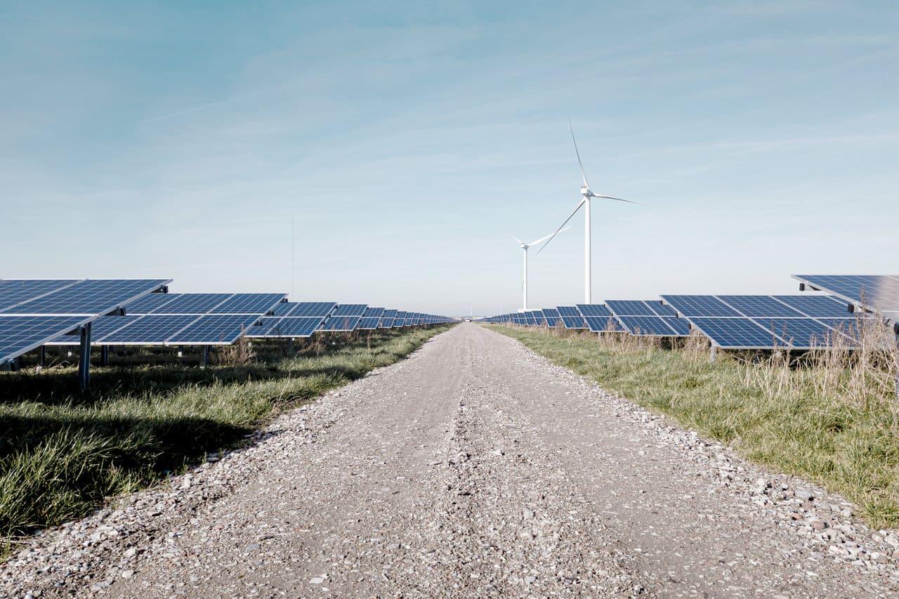 Ein Weg führt durch einen Solarpark. Im Hintergrund sind Windräder zu sehen.