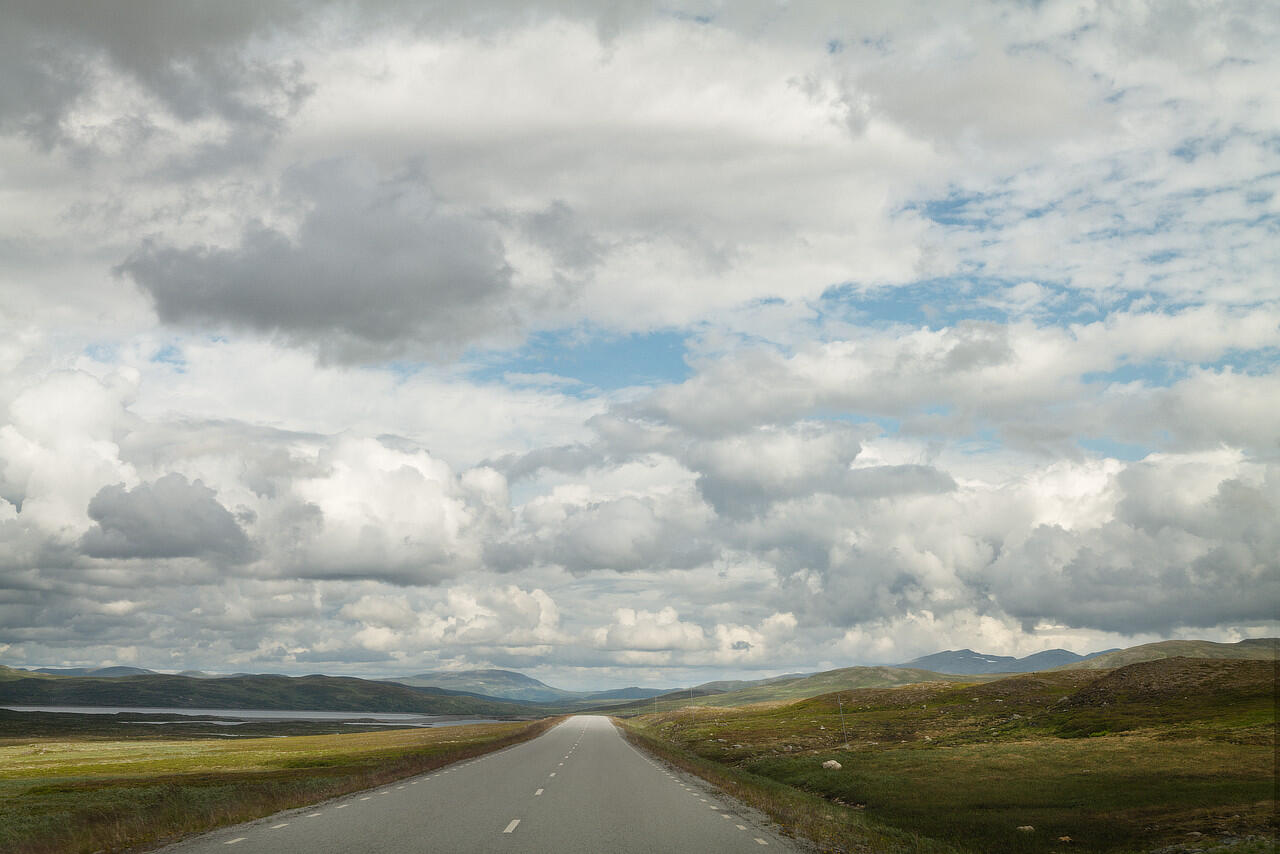 Eine Landstraße führt durch eine grüne Landschaft bis zum Horizont. Der Himmel ist leicht bewölkt.